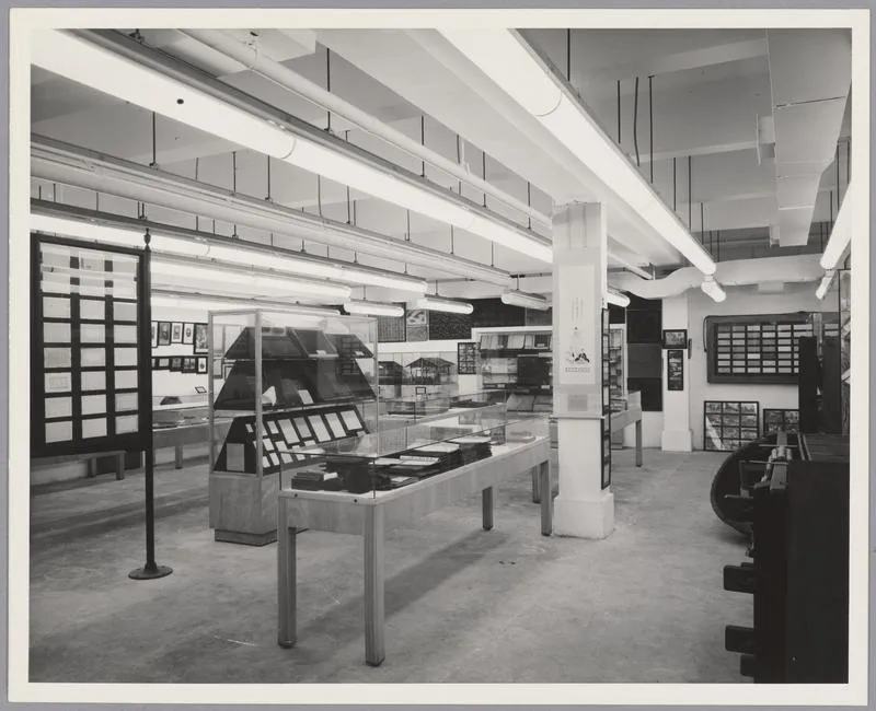 A view of an exhibition in the Dard Hunter Paper Museum while it was at MIT. The exhibit cases in the center of the room display books and prints. On view on the walls surrounding the cases are framed works on paper, and papermaking tools and equipment.